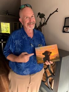 Man standing up holding a copy of Baxter the Highland Unicorn and pointing to it